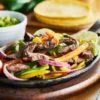 mexican beef fajitas in iron skillet with bell peppers and guacamole on the side